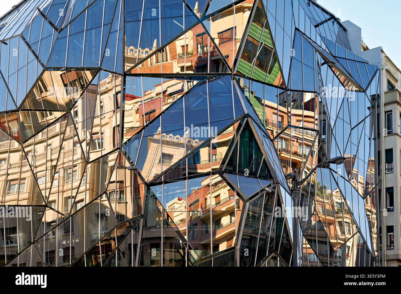 Ein modernes Gebäude in Bilbao spiegelt die umgebende Architektur bei Tageslicht wider. Die Glasfassade zeigt Elemente der Stadt und des Himmels. Stockfoto