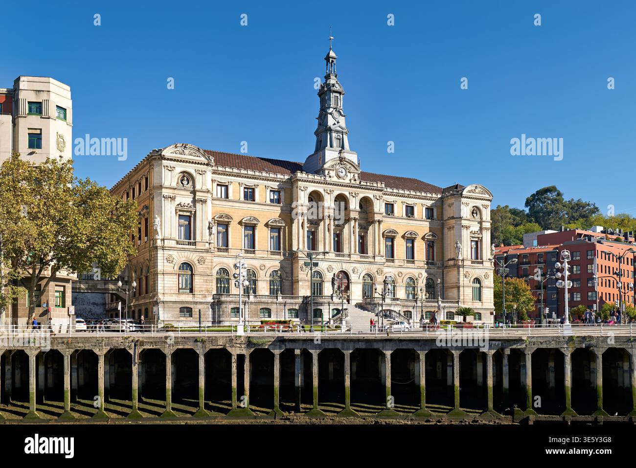 Das Rathaus in Bilbao zeigt seine historische Architektur am Fluss mit Gebäuden und Bäumen, die es unter einem klaren blauen Himmel umgeben. Stockfoto