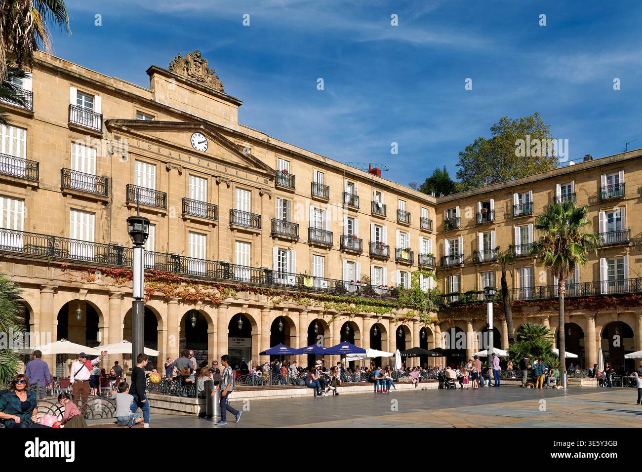 Die Leute spazieren und entspannen sich auf der Plaza Nueva, einem belebten Platz in Bilbao, Spanien. Cafés und Gebäude umgeben die Gegend unter klarem Himmel. Stockfoto
