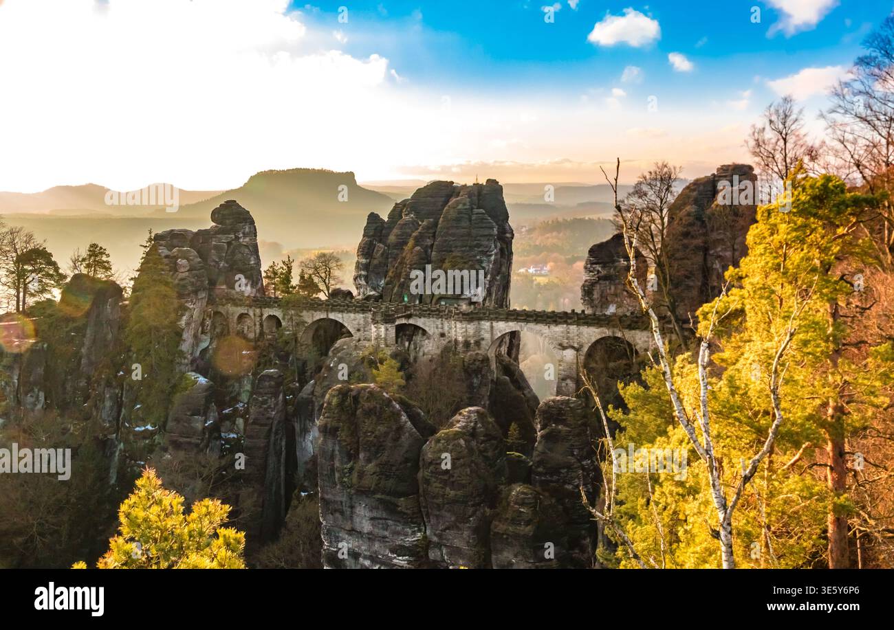 Panoramablick auf den Sonnenaufgang der Bastei-Brücke, die im goldenen Winterlicht leuchtet und über hoch aufragende Sandsteinfelsen gebaut wurde, mit dem entfernten Lilienstein auf dem... Stockfoto