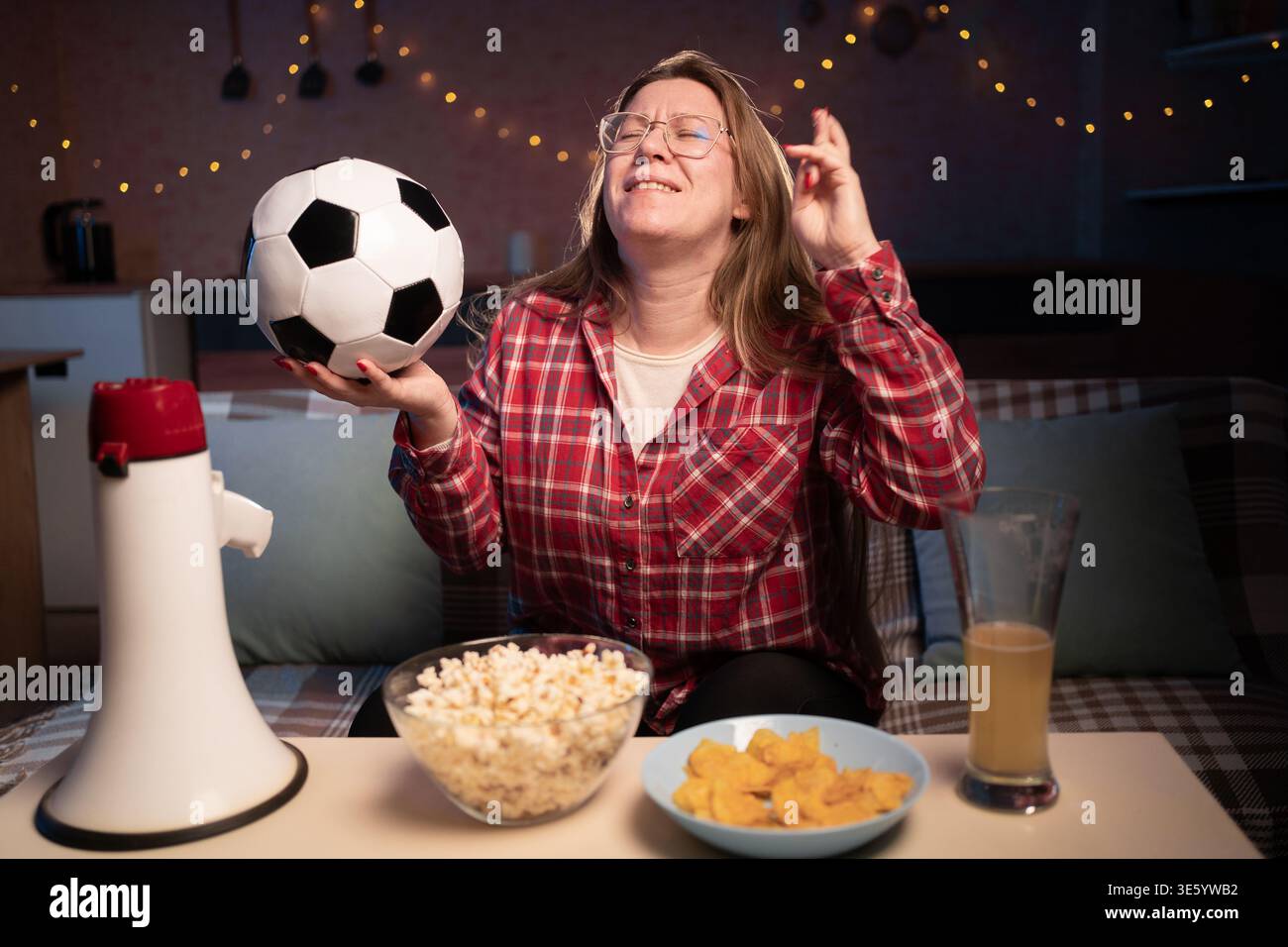 Fußballfan, der zu Hause Fußball ansieht, auf dem Sofa sitzt, mit Bier und Snacks gekreuzt, wünscht und Hoffnung. Sport, le Stockfoto