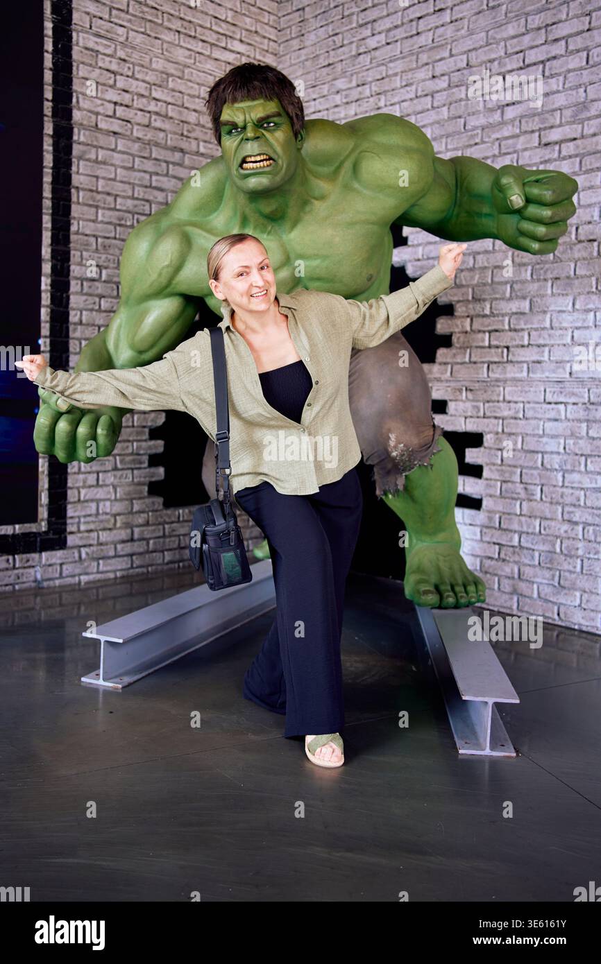 Frau posiert mit einer lebensgroßen Hulk-Statue an einer Ziegelmauer. Lustiger Reisemoment, Fan von Filmcharakteren, Indoor-Entertainment und Freizeitkonzept. Stockfoto