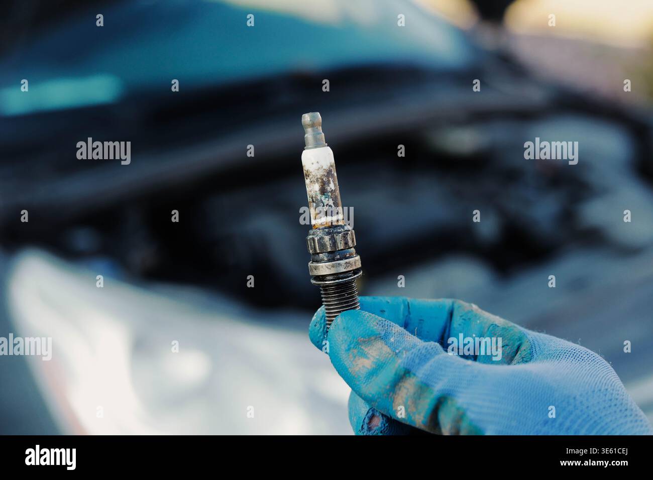 Der Automechaniker hält die verbrannte Zündkerze in der Hand nah mit dem Auto im Hintergrund. Servicekonzept für Fahrzeugwartung, Motorausfall und Reparatur. Stockfoto