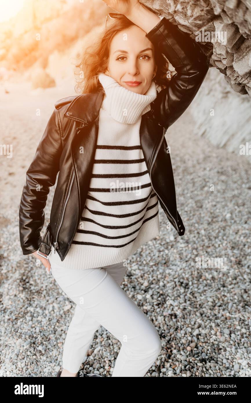 Frau Strandmode elegante Frau mit Lederjacke und gestreiftem Pullover posiert an einem Kieselstrand unter warmem Sonnenlicht Stockfoto