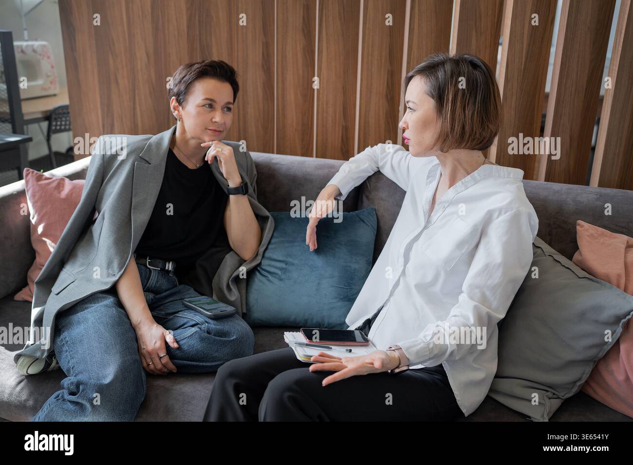 Frauen auf der Arbeit lachen und reden zusammen auf dem Sofa in einem hellen Büro. Lachen, reden, Teamarbeit, Büro, Geschäftsfrauen, freundlich. Stockfoto
