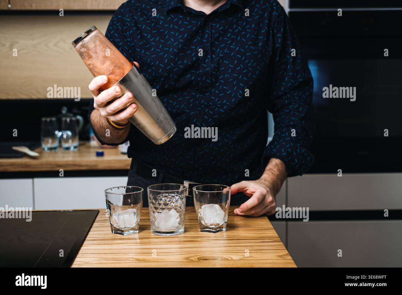 Mann schüttelt Metall Cocktail Shaker über drei Gläser gefüllt mit Eis auf Holztheke. Professioneller Barkeeper, Cocktailmixologie, Craft Drinks Stockfoto