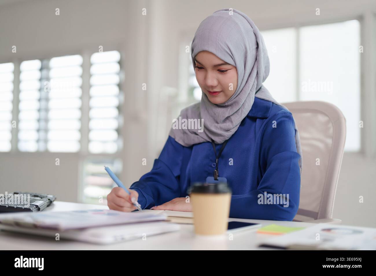 Fleißige asiatische muslimische Frau, die sich auf Papierkram konzentriert. Professionelle Lernerin führt Datenanalysen durch und schreibt Notizen zu Dokumenten am Schreibtisch. Stockfoto