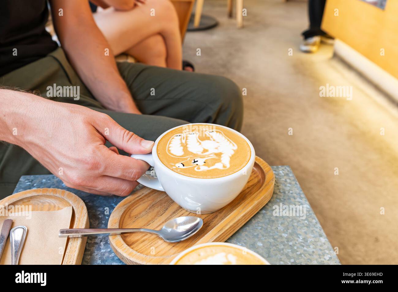 Mann Hand hält eine Tasse Cappuccino mit detaillierter Latte Art in Form eines Bären, während er im Café sitzt und auf Holztablett serviert wird. Nahaufnahme. Kaffeegetränk und Stockfoto