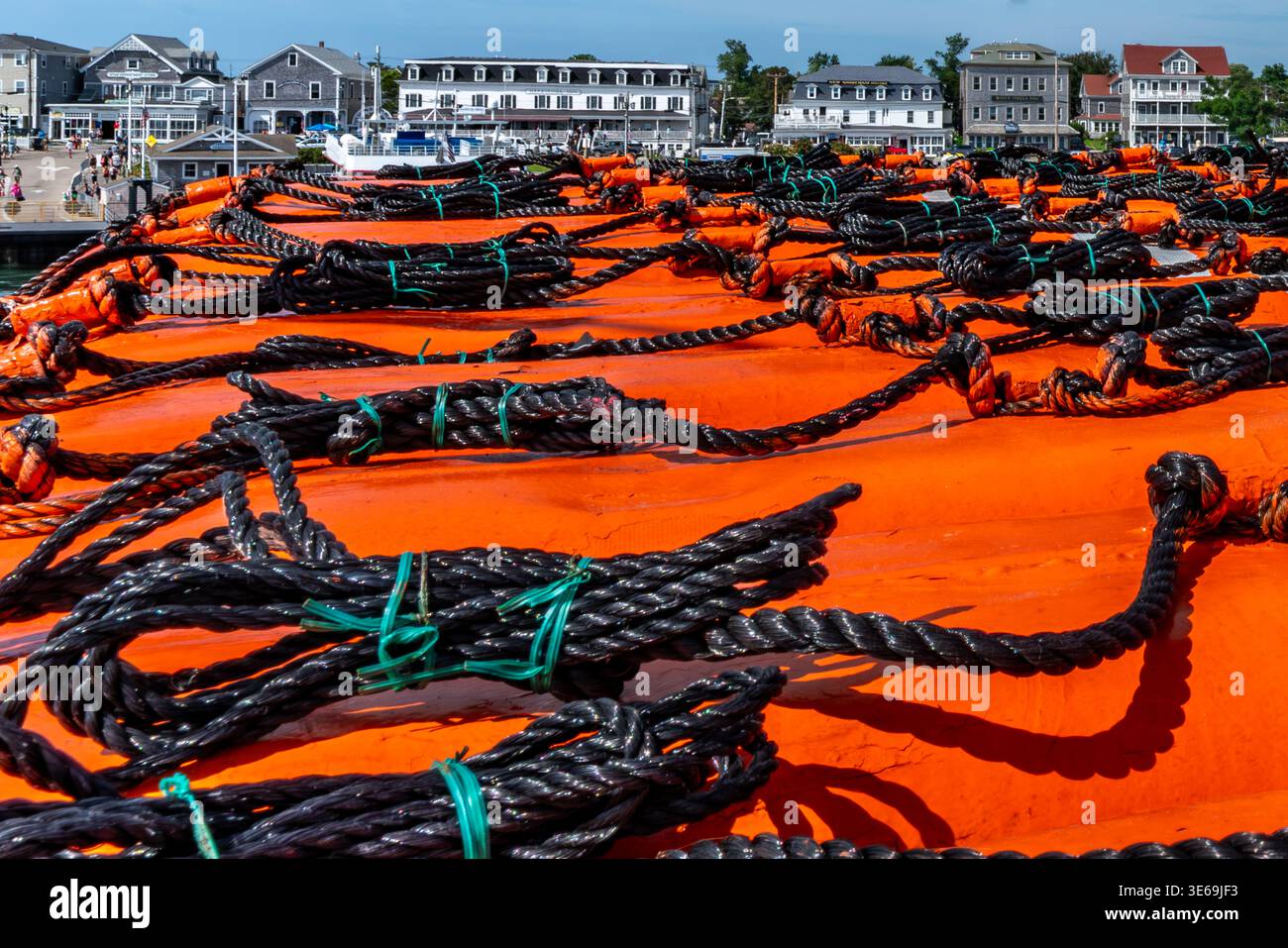 Im Vordergrund befinden sich schwarze Seile mit blaugrünen Bändern auf einer leuchtend orangefarbenen Oberfläche, mit einer malerischen Küstenstadt und einem Hafen im Hintergrund. Stockfoto