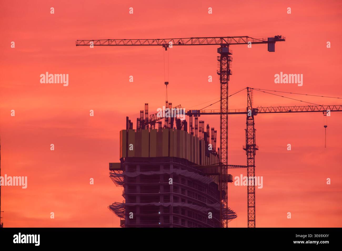 Hochhaus im Bau mit Turmkränen Silhouetten vor dem roten Sonnenuntergang. Tragender Rahmen und Hebezeug in Betrieb Stockfoto