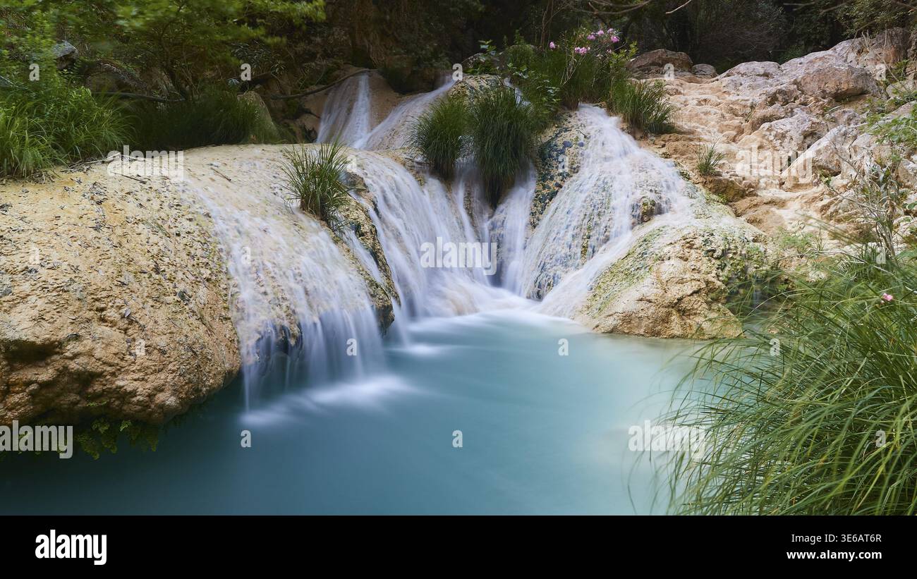 Ein kleiner Wasserfall fließt über Felsen in einen klaren türkisblauen Pool, der von Pflanzen, Wasserfällen, Teichen, Polylimnio, Peloponnes, Griechenland Stockfoto