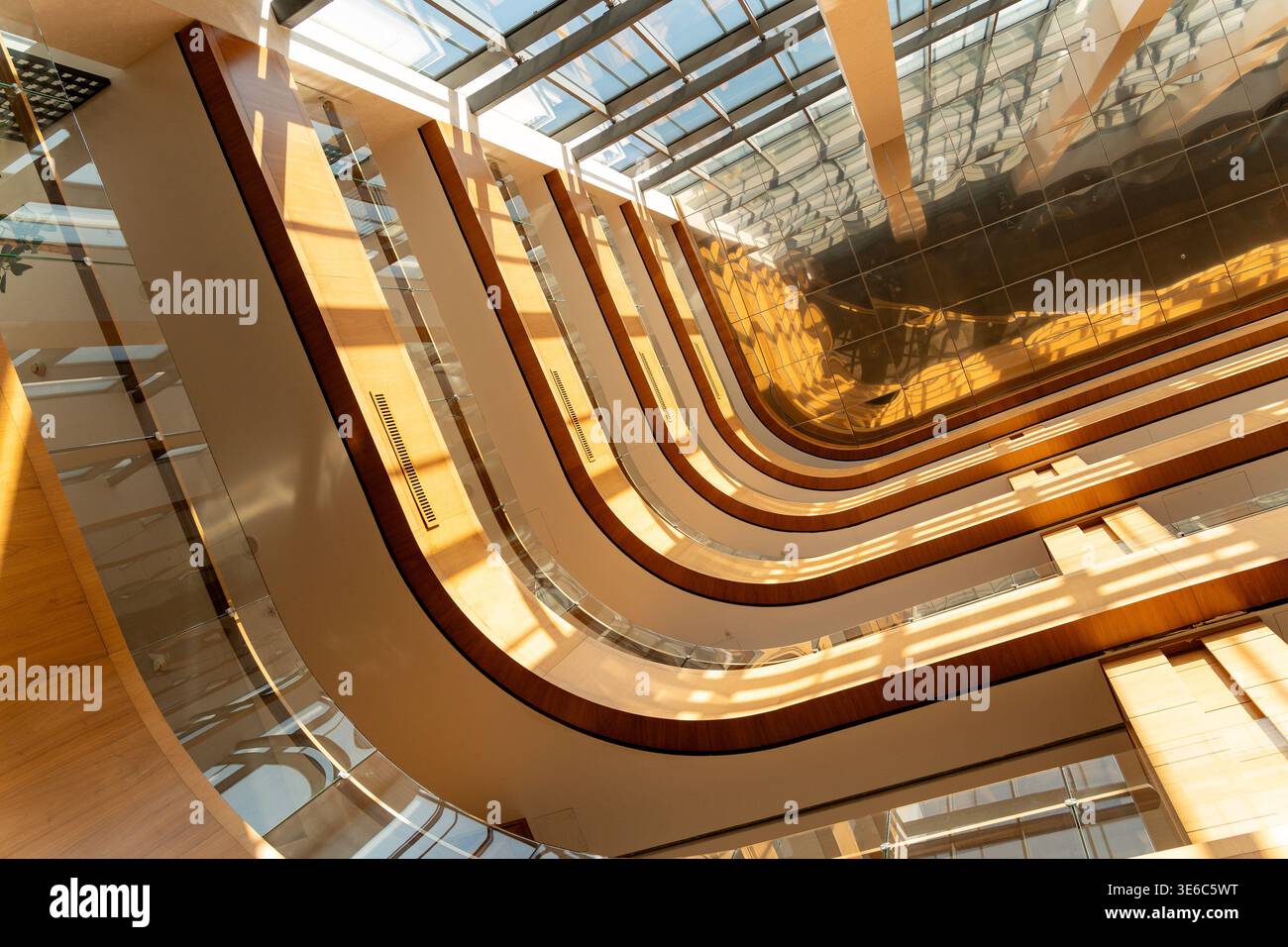Modernes Atrium mit geschwungenen Balkonen, Glasgeländern, Holzakzenten und einer Glasdecke, die Sonnenlicht und Schatten auf reflektierenden Oberflächen ausstrahlt. Stockfoto