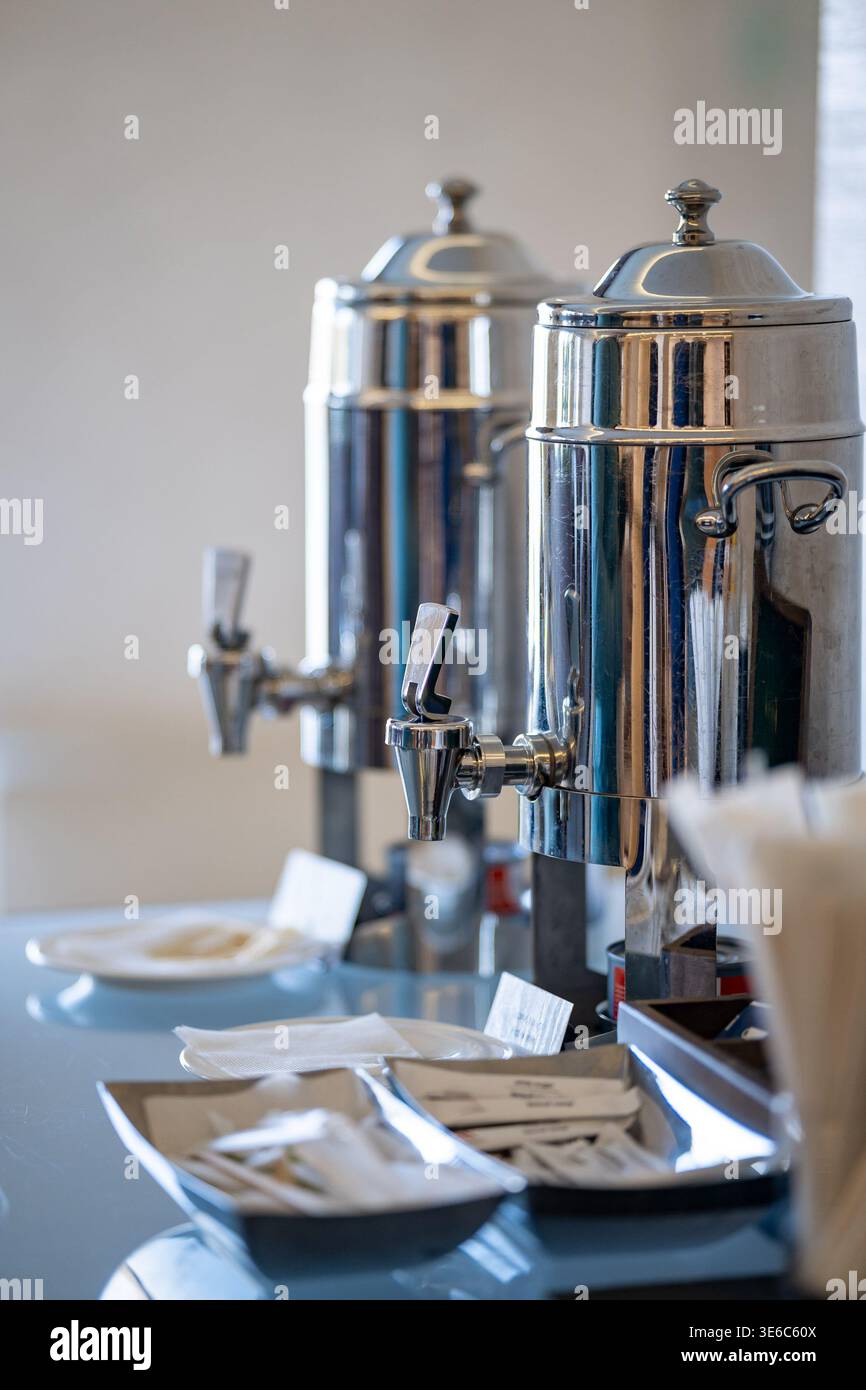 Selbstbedienungs-Kaffeestation mit glänzenden Edelstahl-Getränkeurnen, Zuckerpackungen, Servietten und Keksen auf einem Tisch. Stockfoto