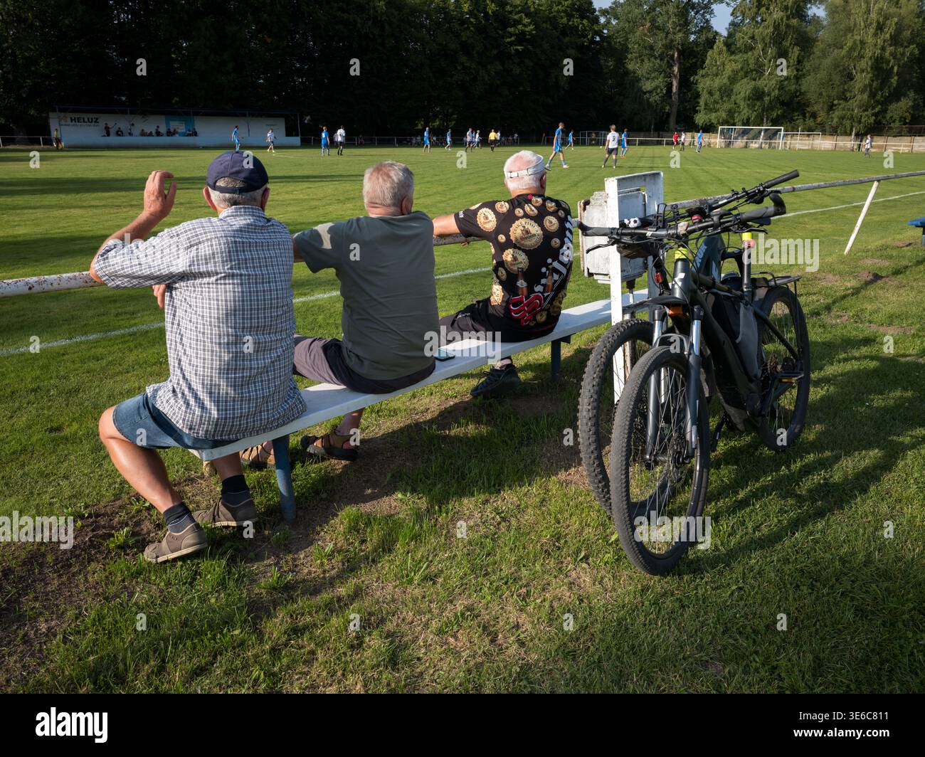Ältere Männer sitzen auf einer Bank und beobachten ein lokales Fußballspiel mit ihren Fahrrädern in der Nähe Stockfoto