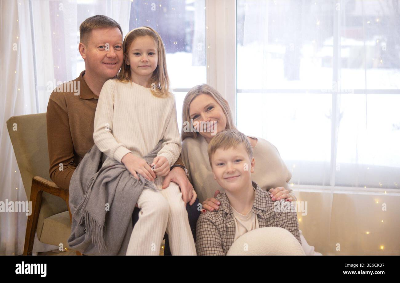 Eine vierköpfige Familie, darunter zwei Kinder, posiert in Winterkleidung drinnen, mit einer verschneiten Szene vor dem Fenster und sanfter Beleuchtung, die den gemütlichen Lärm unterstreicht Stockfoto