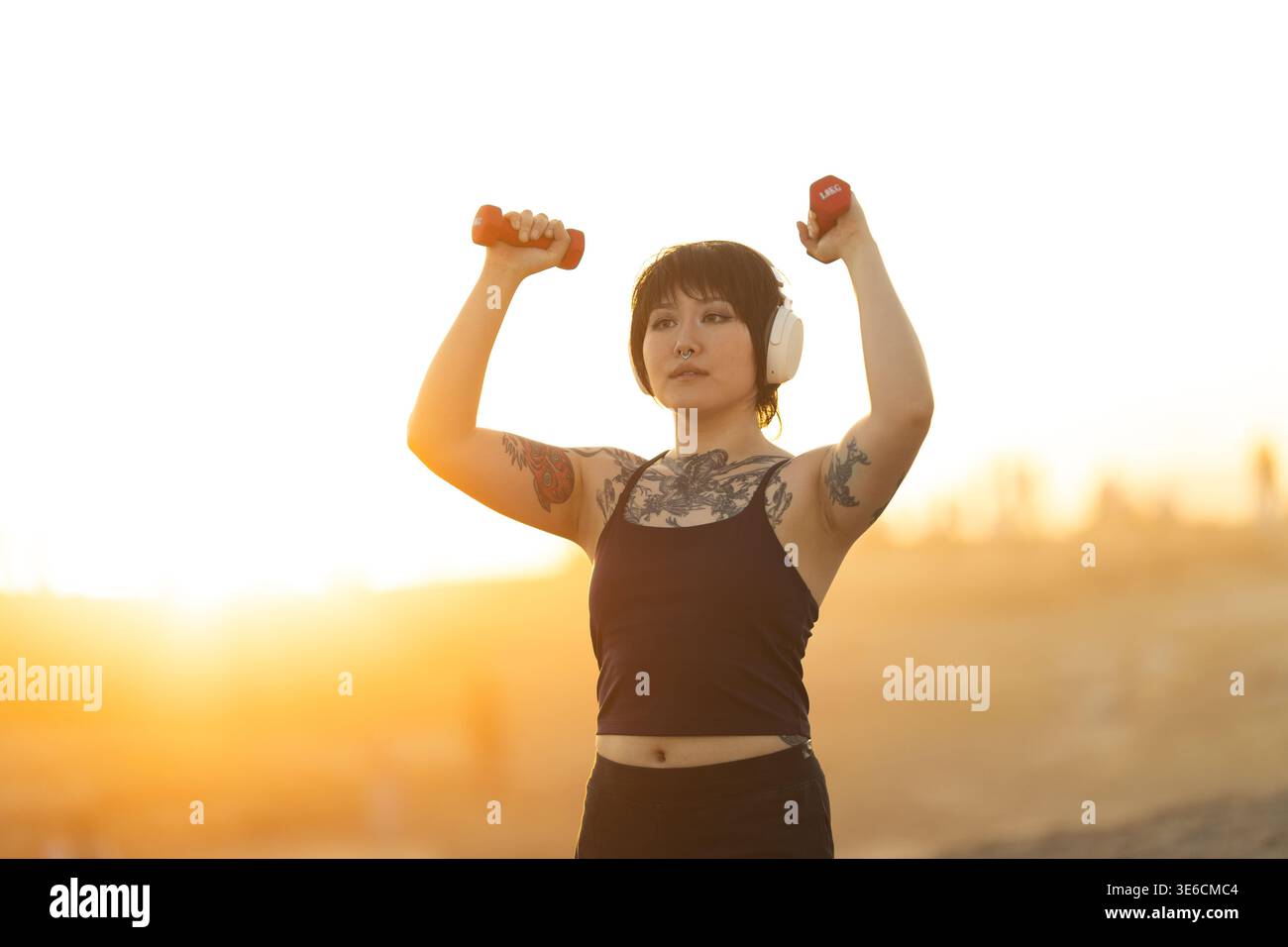 Frau, die während der goldenen Stunde ein Outdoor-Workout mit Kurzhanteln mit Kopfhörern durchführt Stockfoto