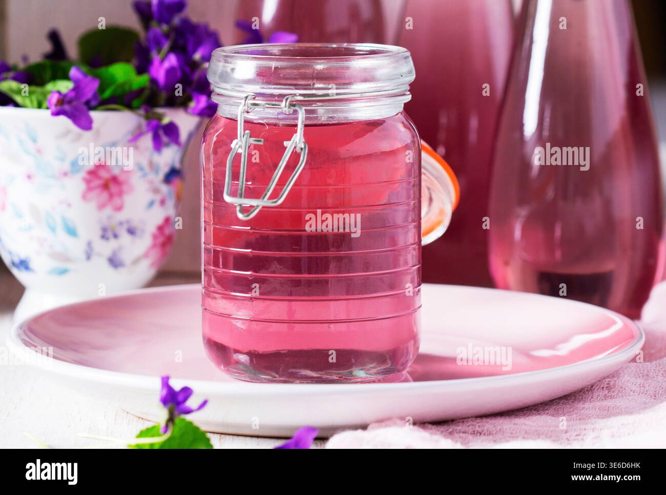 Violetter Sirup in einem Glasgefäß, kombiniert mit einem Bouquet frischer Veilchen, kreiert eine zarte florale Komposition mit weichen Pastelltönen und natürlichem Licht. Stockfoto
