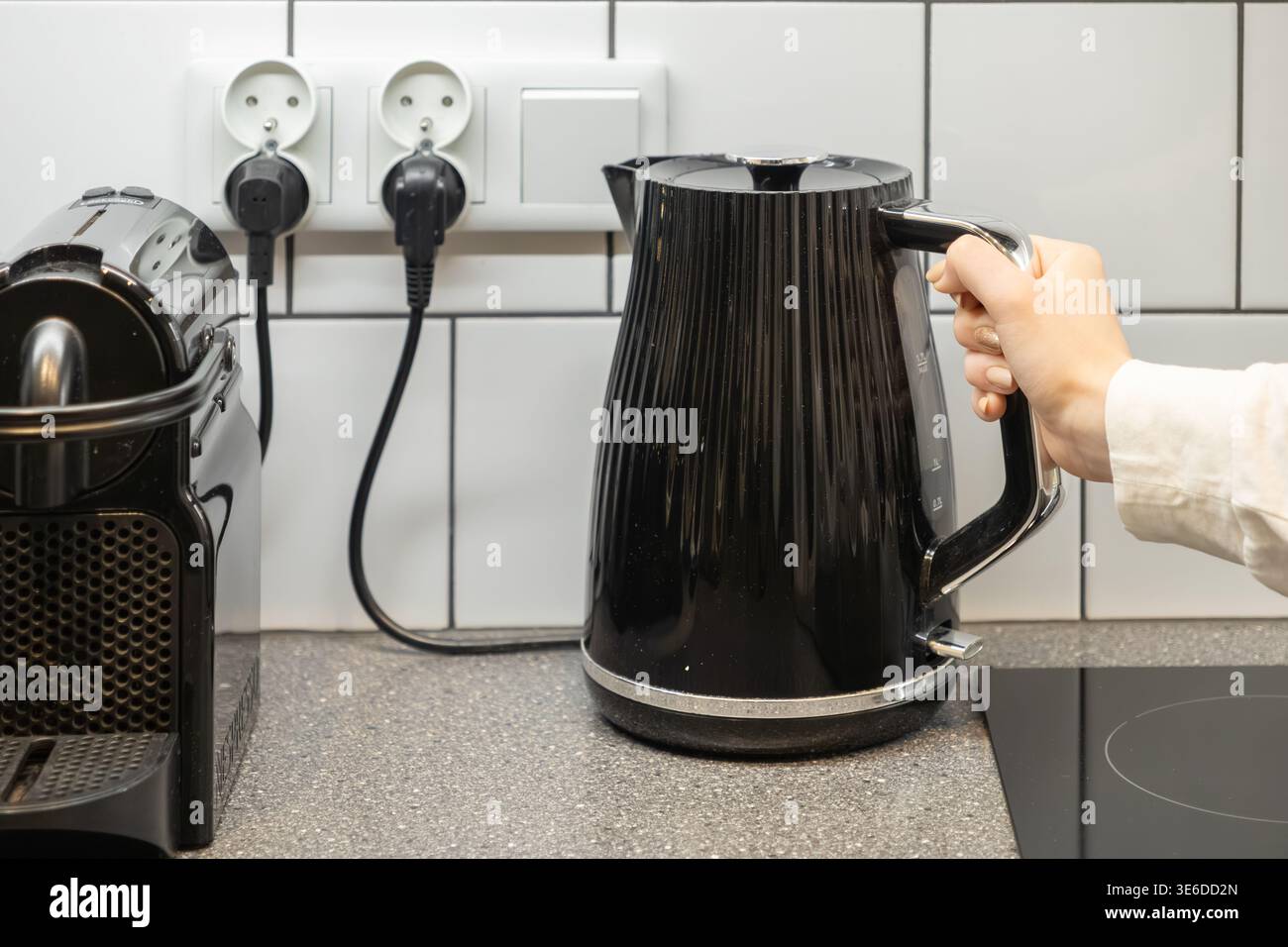 Detaillierte Ansicht eines handgreifenden, eleganten schwarzen Wasserkochers in der Nähe einer kompakten Kaffeemaschine, der Effizienz und Komfort in einer modernen Wohnung symbolisiert Stockfoto