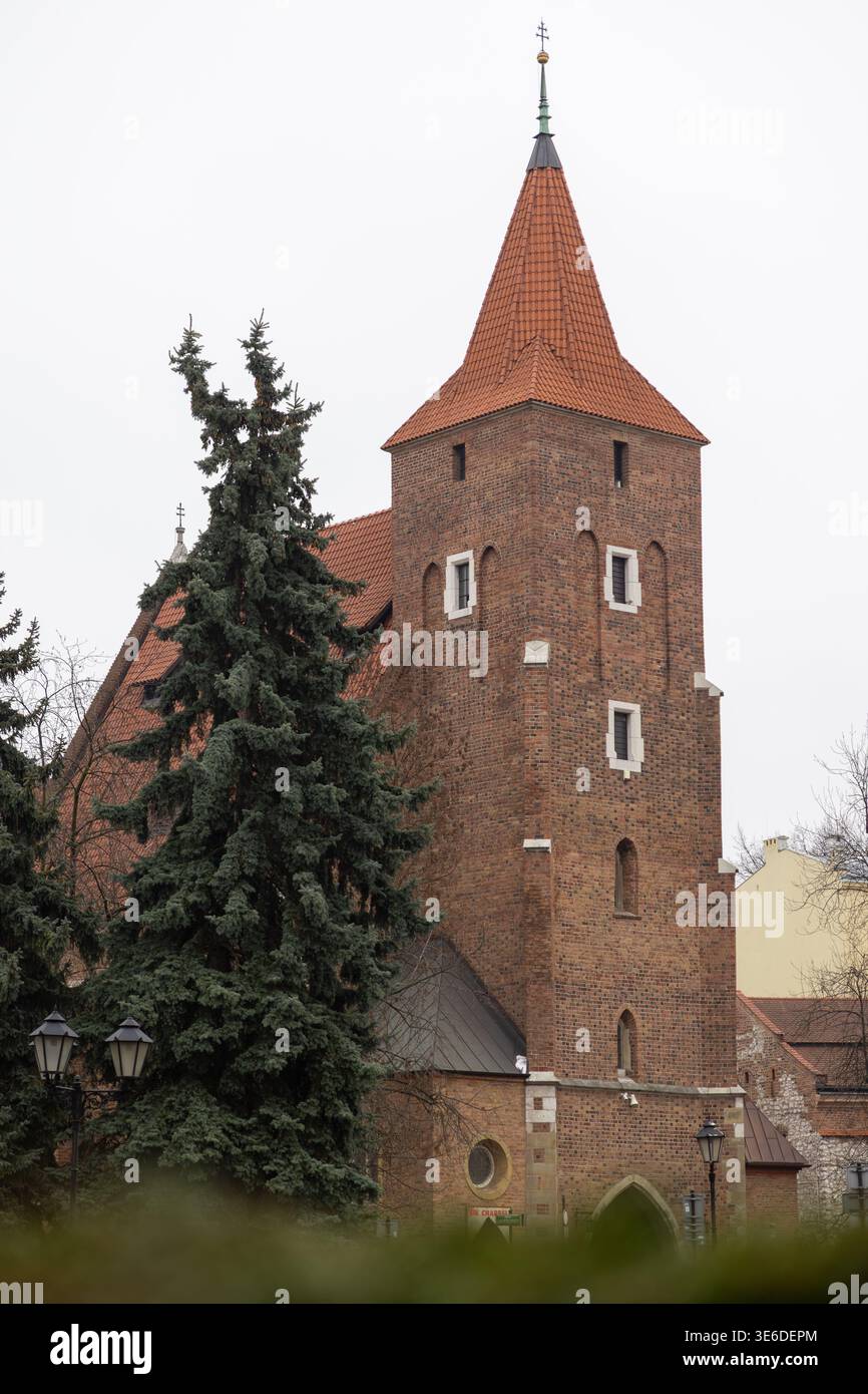 Krakau, Polen - 1. Dezember 2025: Europäischer Kirchturm, roter Backstein, Ziegeldach, Spitzturm, grüne Tannen, das historische religiöse Archit darstellt Stockfoto