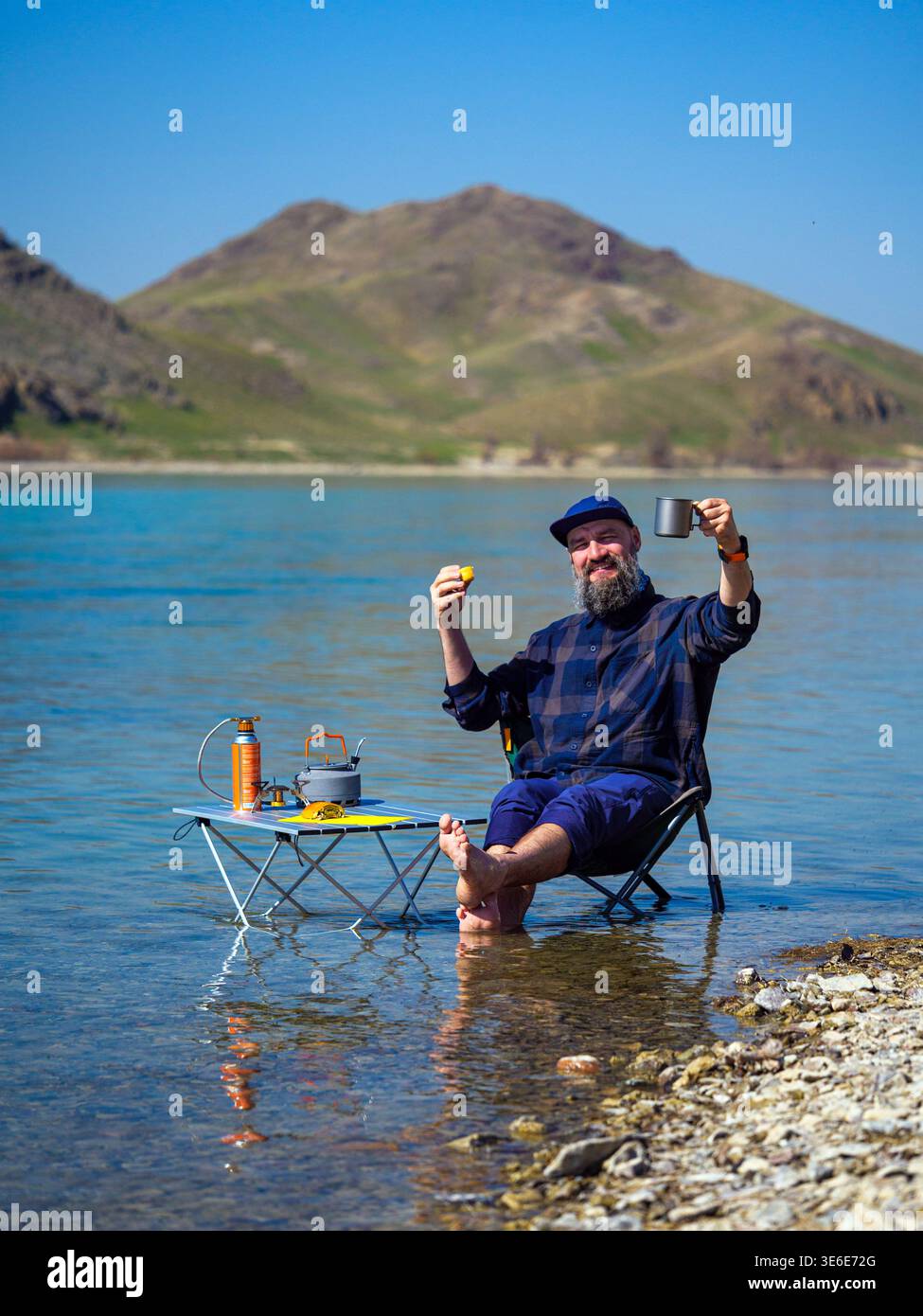 Ein bärtiger Mann entspannt sich im Wasser am Ufer. Ein fröhlicher Mann lagert im Fluss, sitzt auf einem Klappstuhl und trinkt Kaffee Stockfoto