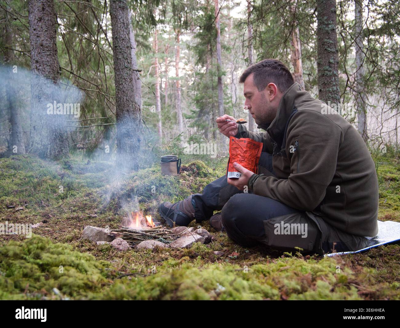 Mann isst Überlebensmahlzeit am Lagerfeuer im Wald Schwedens Stockfoto