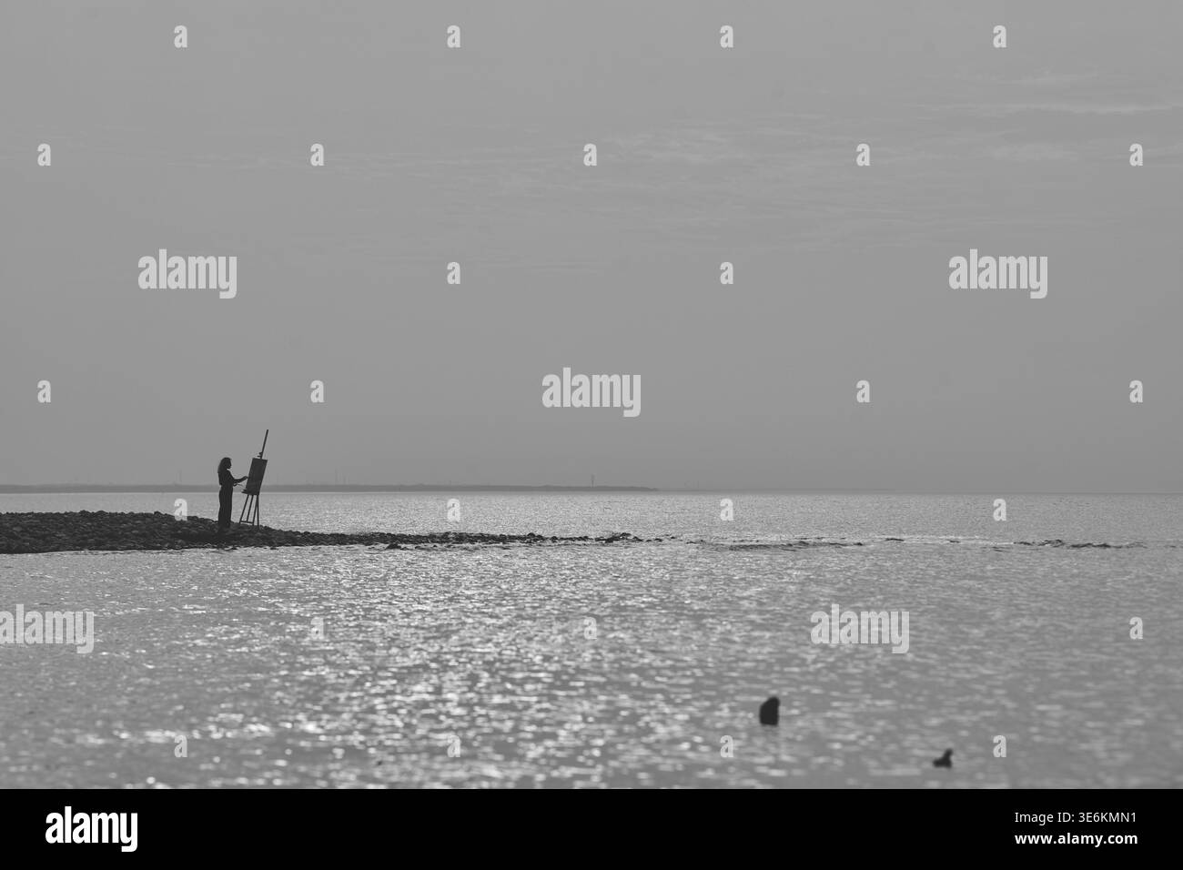 Silhouette des Stehens und Malens von Erwachsenen auf der Staffelei an der felsigen Küste bei Sonnenuntergang, mit glitzerndem Wasser und ruhigem Horizont in Grautönen Stockfoto