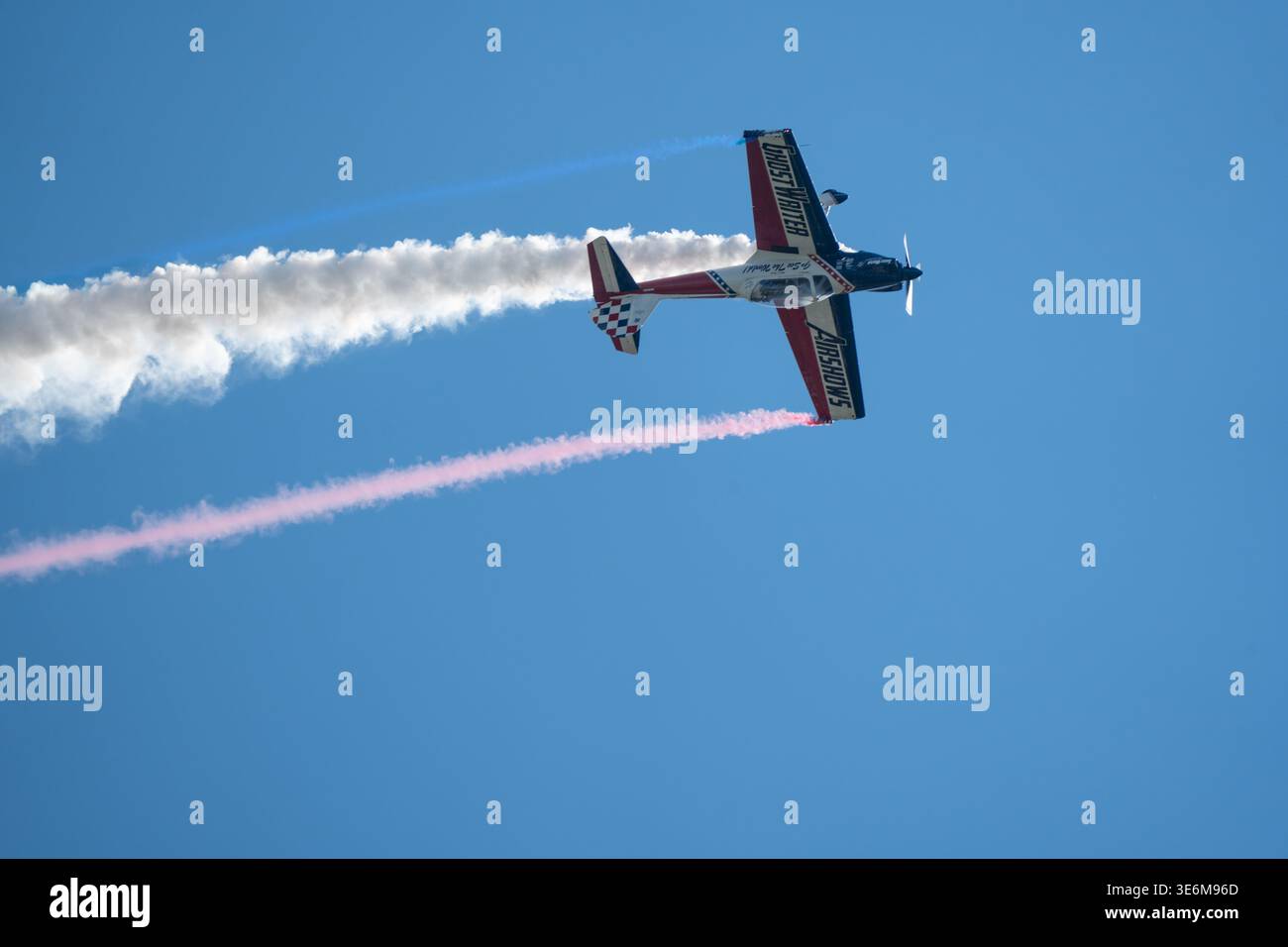 Ein Hochgeschwindigkeits-Kunstflugzeug, das während einer Flugshow farbenfrohe Rauchpfade vor einem klaren blauen Himmel erzeugt und Präzisionsfliegen zeigt Stockfoto