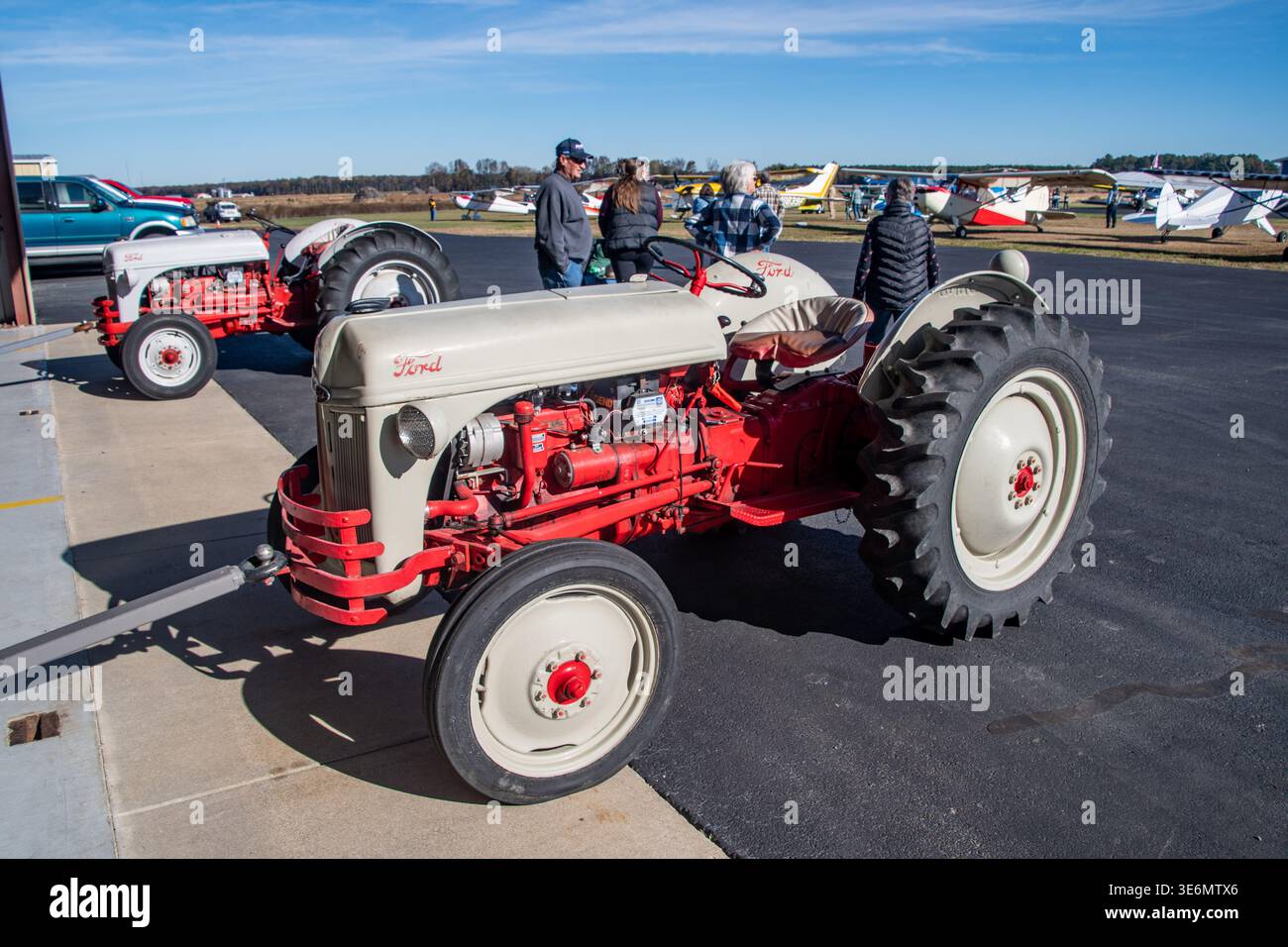 Roter und cremefarbener Ford-Traktor, der auf einem kleinen Flughafenparkplatz mit leichten Flugzeugen und Zuschauern in der Nähe geparkt wurde. Heller, sonniger Himmel, ländlicher Flugplatz Stockfoto