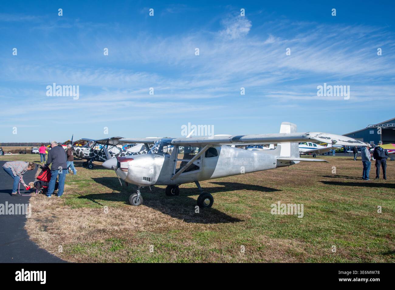 Ein kleines Aluminiumflugzeug sitzt auf dem Gras neben einem asphaltierten Rollweg auf einem belebten Flugplatz, umgeben von Vintage-Leichtflugzeugen, Zuschauern und Hangars Stockfoto
