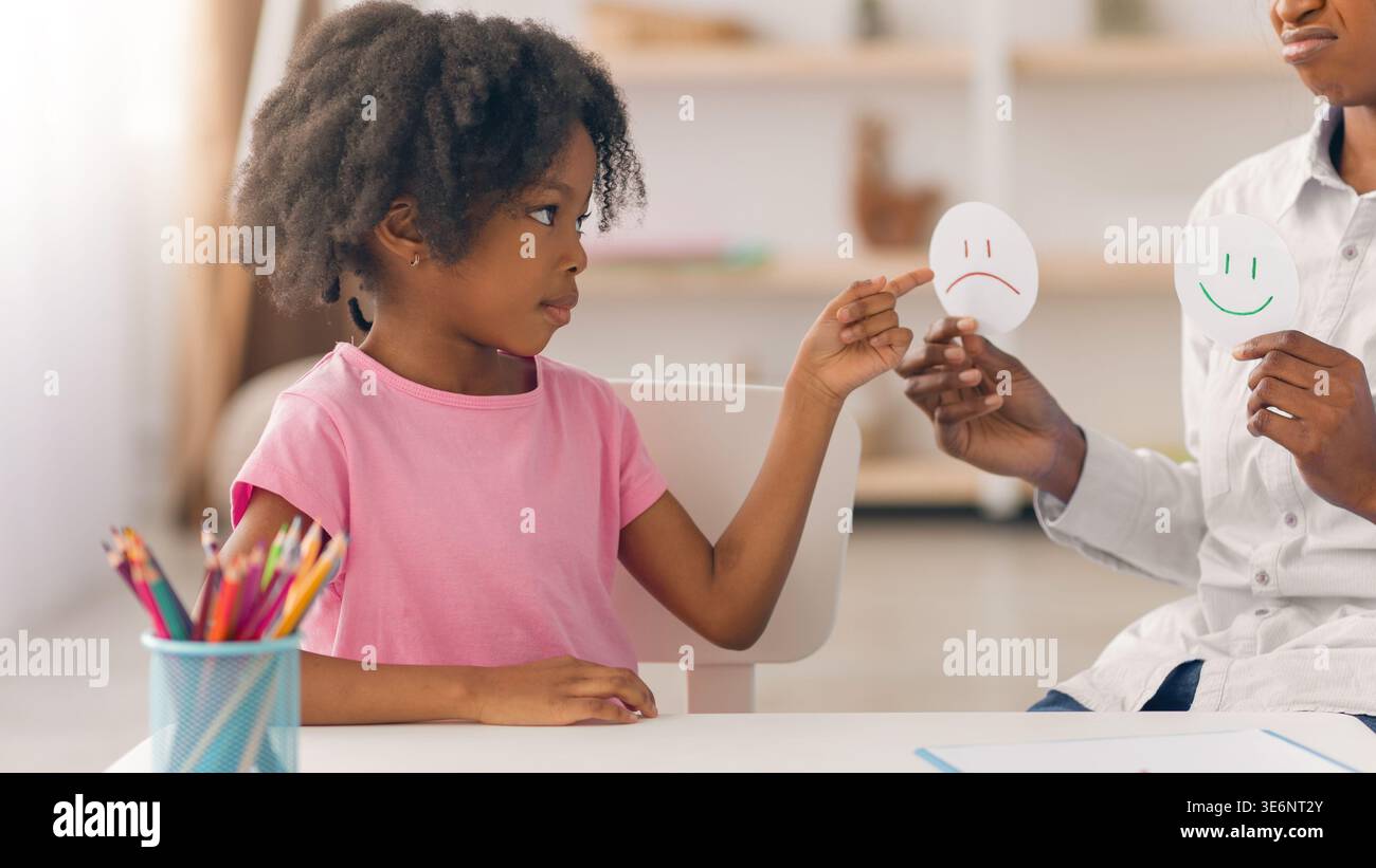 Kind drückt Gefühle mit Smiley Gesichtern aus, während er an einem Tisch in einem hellen Raum sitzt Stockfoto