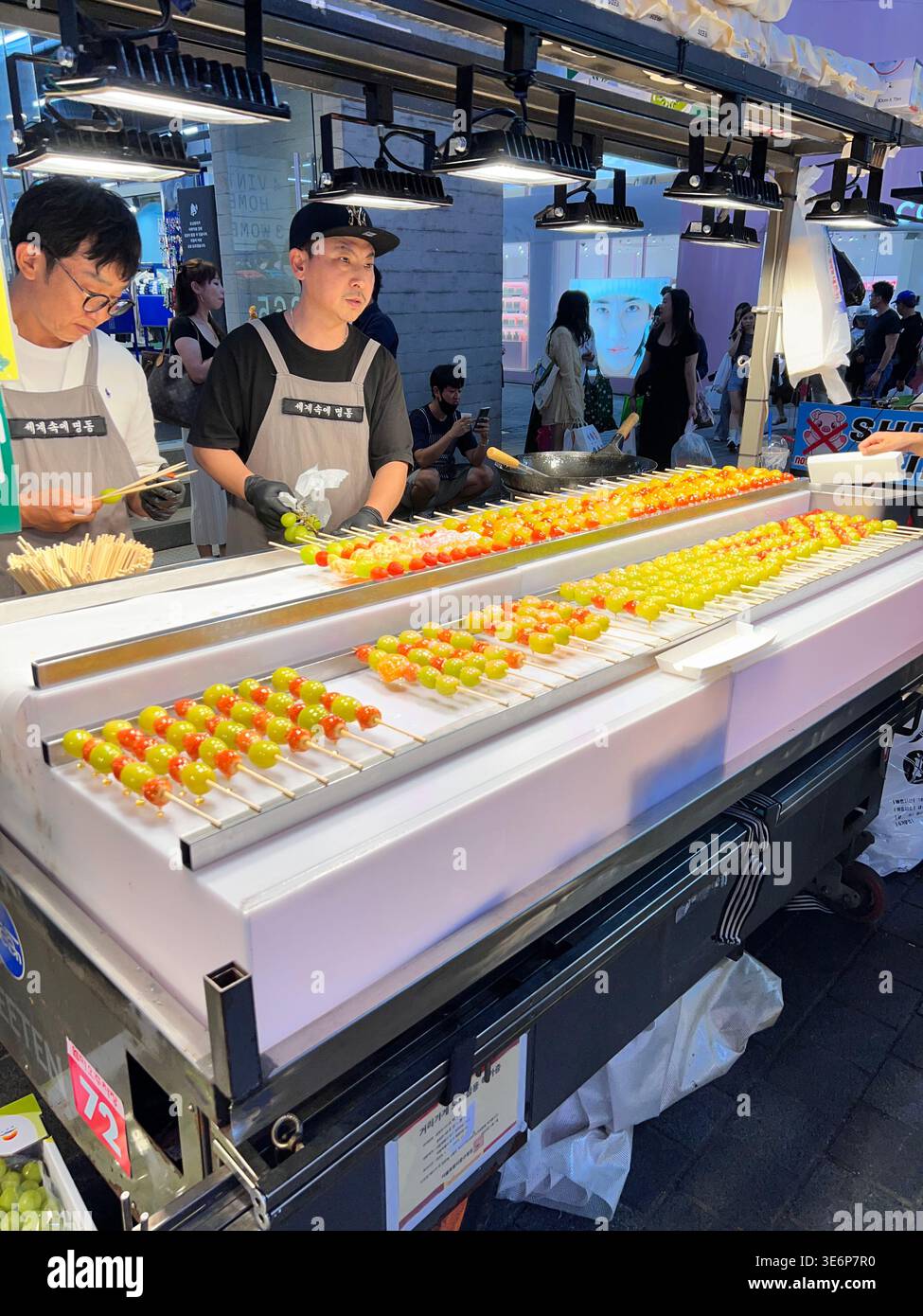Verkäufer, der kandierte Obstspieße an einem Nachtmarkt in Myeong-dong, Seoul, Südkorea, zubereitet. - Smartphone-aufgenommenes Stockfoto