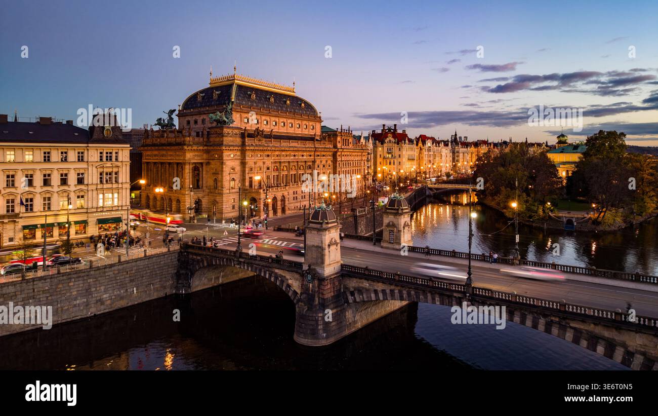 Das Nationaltheater steht am Ufer in Prag, beleuchtet von Abendlichtern. Der Verkehr fließt über die nahe gelegene Brücke, während die Stadt in die Nacht übergeht. Stockfoto