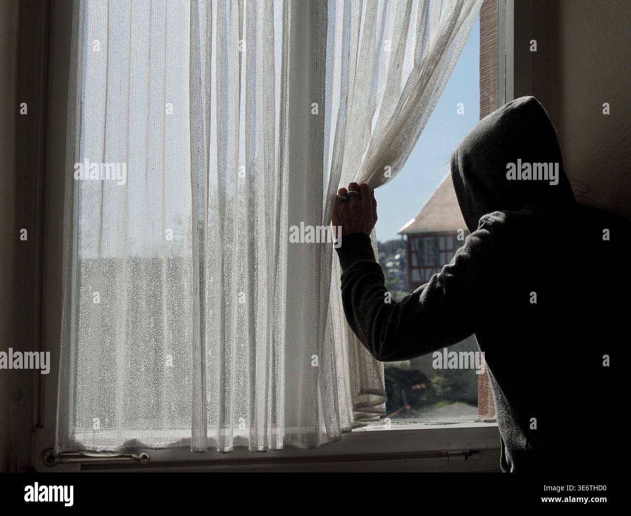 Eine Figur in einem Kapuzenpullover schiebt den Vorhang zur Seite und blickt aus einem Fenster. Stockfoto