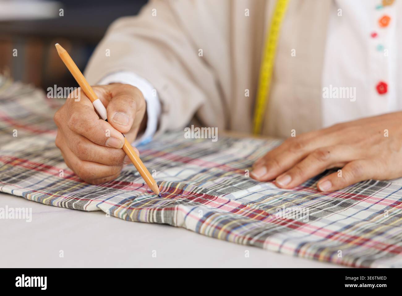 Eine Nahaufnahme eines professionellen Schneiders, der in einem kreativen Modestudio präzise Maße auf einem Stück karierten Stoff mit einem Bleistift markiert. Stockfoto