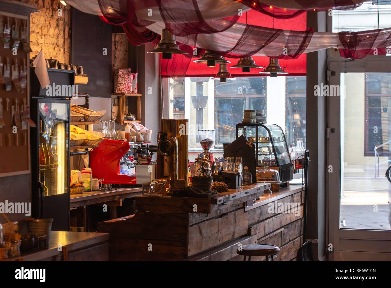 Ein gemütliches Café mit roter Espressomaschine, Gläsern, Desserts in einer Vitrine und warmer Beleuchtung mit Fensterblick im Hintergrund. Stockfoto