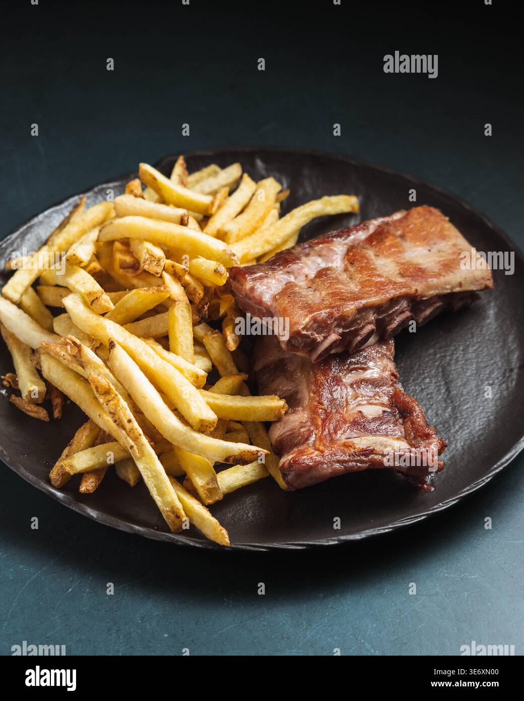 Gegrillte Rippchen sitzen neben goldenen Pommes Frites auf dunklem Teller. Textur und Farbe lassen eine zarte, aromatische Zubereitung vermuten. Einfache, auffällige Präsentation betont das Hören Stockfoto