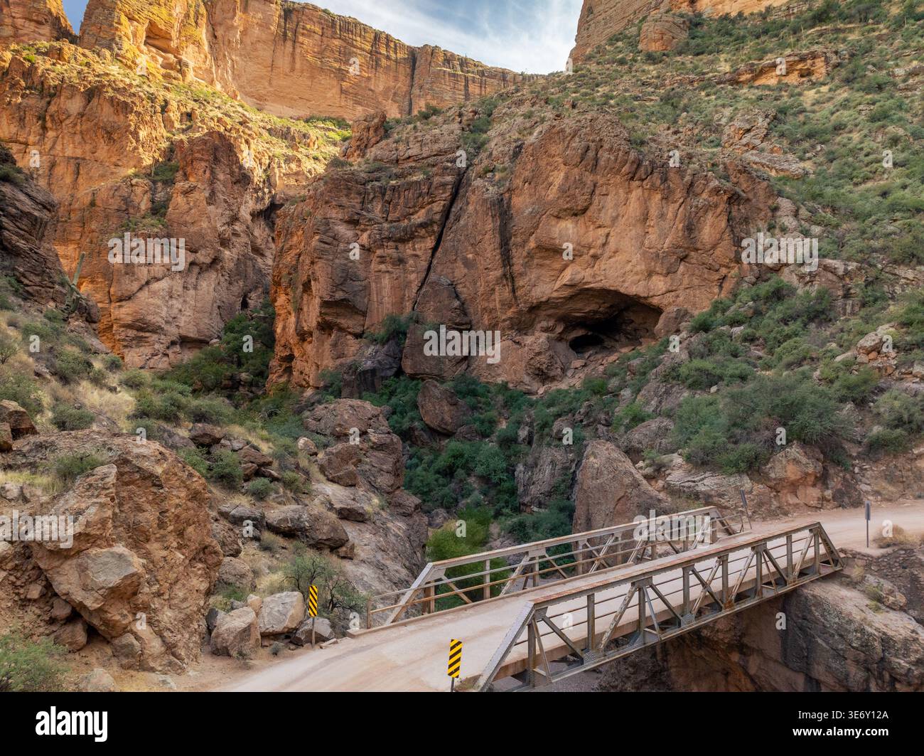 Eine Metallbrücke überquert den engen Canyon auf dem Apache Trail mit massiven roten Felsklippen und Wüstenvegetation in der Nähe von Phoenix Arizona unter teilweise bewölktem Himmel. Stockfoto