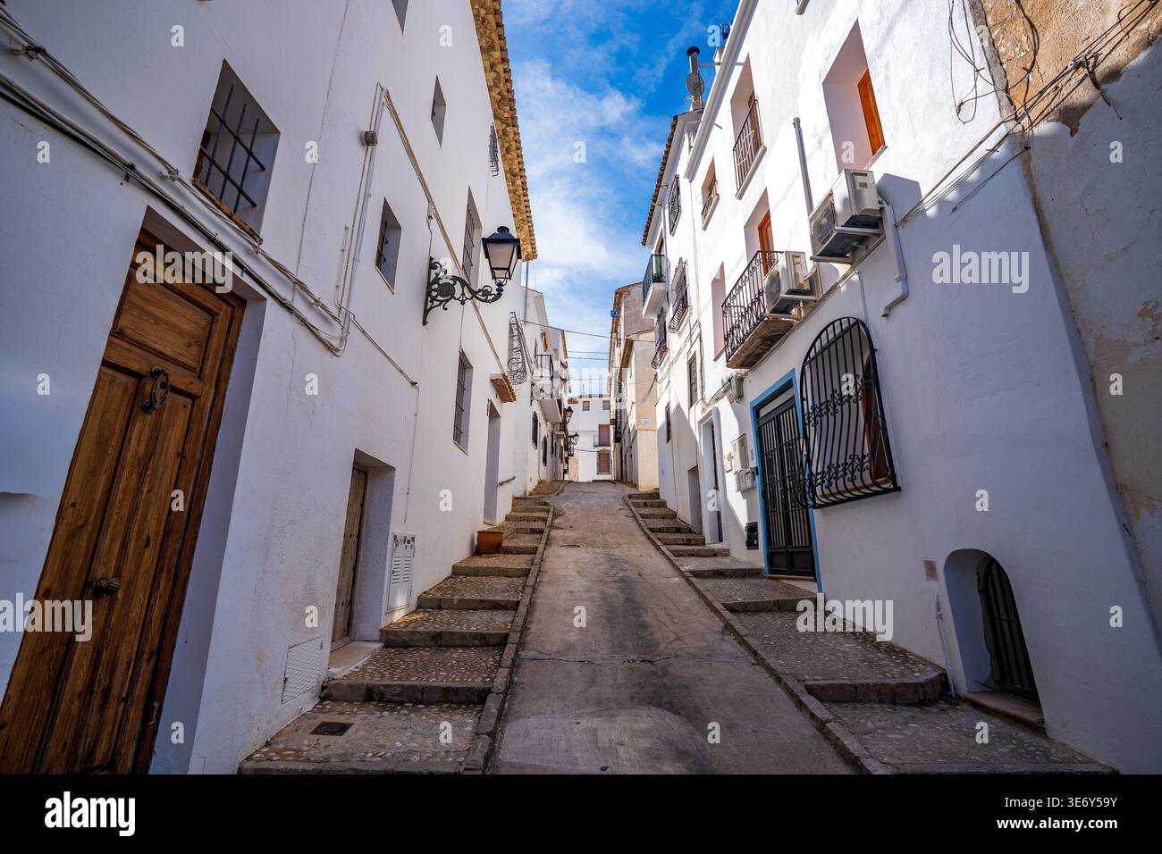 Eine malerische Kopfsteinpflasterallee gesäumt von hellen weißen mediterranen Häusern unter einem klaren blauen Himmel in Altea. Die zentrale Perspektive führt das Auge nach oben Stockfoto