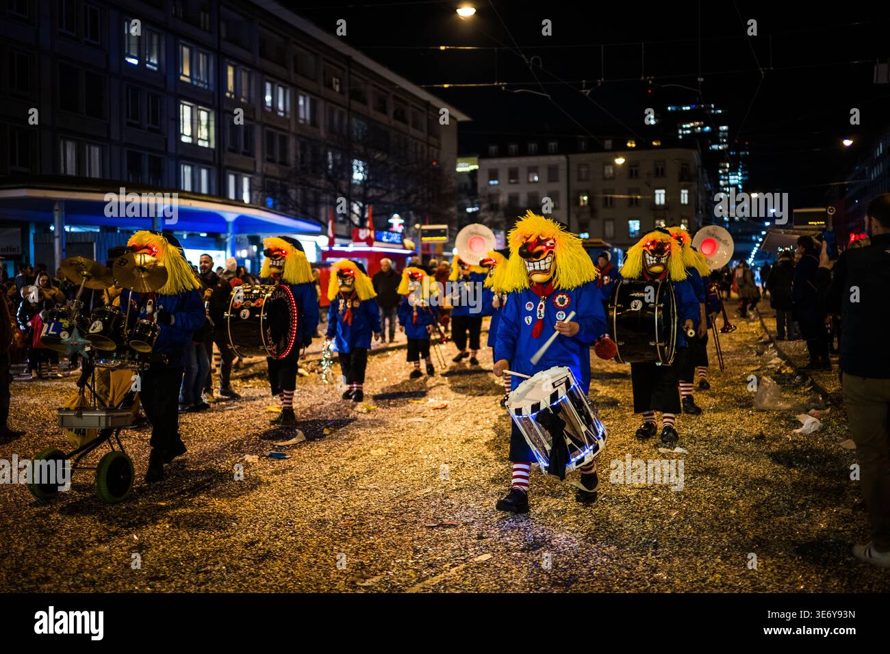 Basel, Schweiz - 10. März 2025: Blick auf eine pulsierende Parade mit maskierten Musikern in bunten Kostümen, die durch die mit Konfetti übersäten Straßen A marschieren Stockfoto