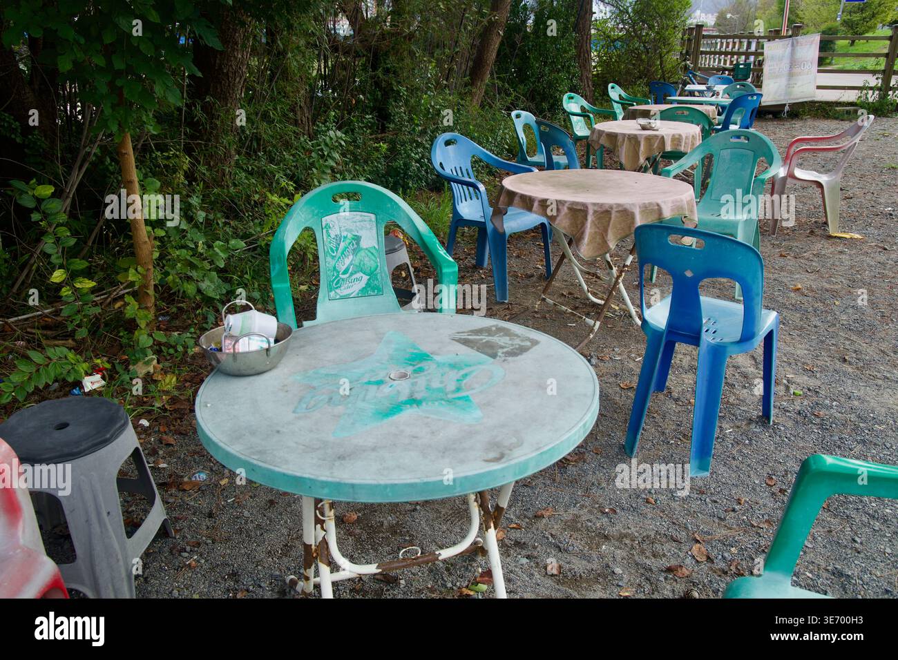 Plastiktische und nicht übereinstimmende Stühle sitzen vor einer Kaffee- und Radtour-Haltestelle in der Nähe des Seonbawi Rock, beschattet von Bäumen entlang des Taehwa River in Guyeong- Stockfoto