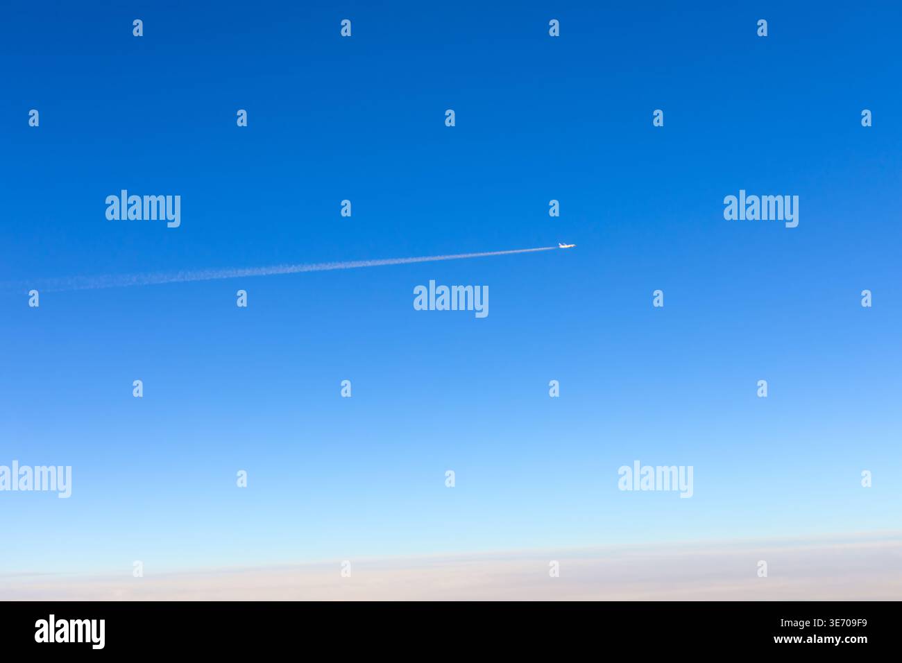 Klarer blauer Himmel mit einem weißen Jet, der von links nach rechts hochfliegt und einen langen, dünnen weißen Kondensstreifen hinterlässt. Eine schwache, trübe Schicht erscheint an der Stelle Stockfoto