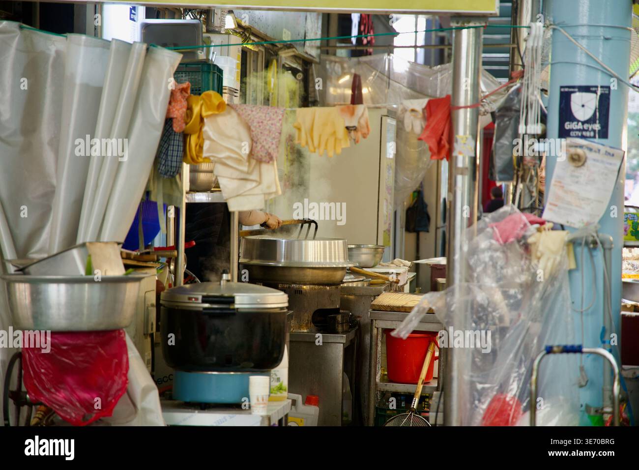 Dampf steigt aus einer funktionierenden Küche im Eonyang Alps Market, wo Plastikvorhänge, Hängetücher und Metalltöpfe das Innere des Verkaufsstandes bevölkern. Stockfoto