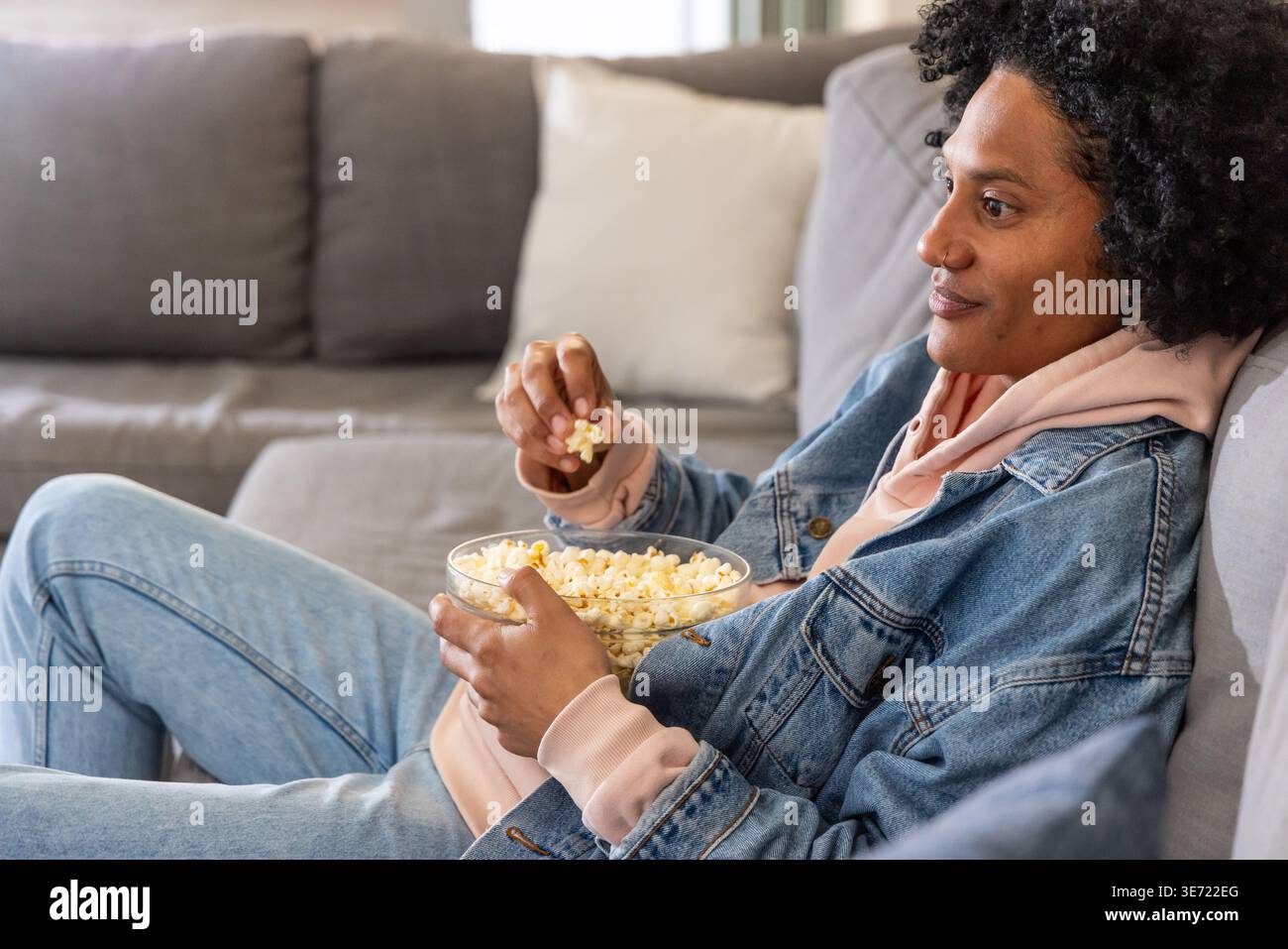 Afroamerikanerin, die sich zu Hause auf dem Sofa in Denim liegend, Glasschale Popcorn, Kopierraum. Gemütlich, Zuhause, Entspannung, lässig, weich, natürlich, Neutr Stockfoto