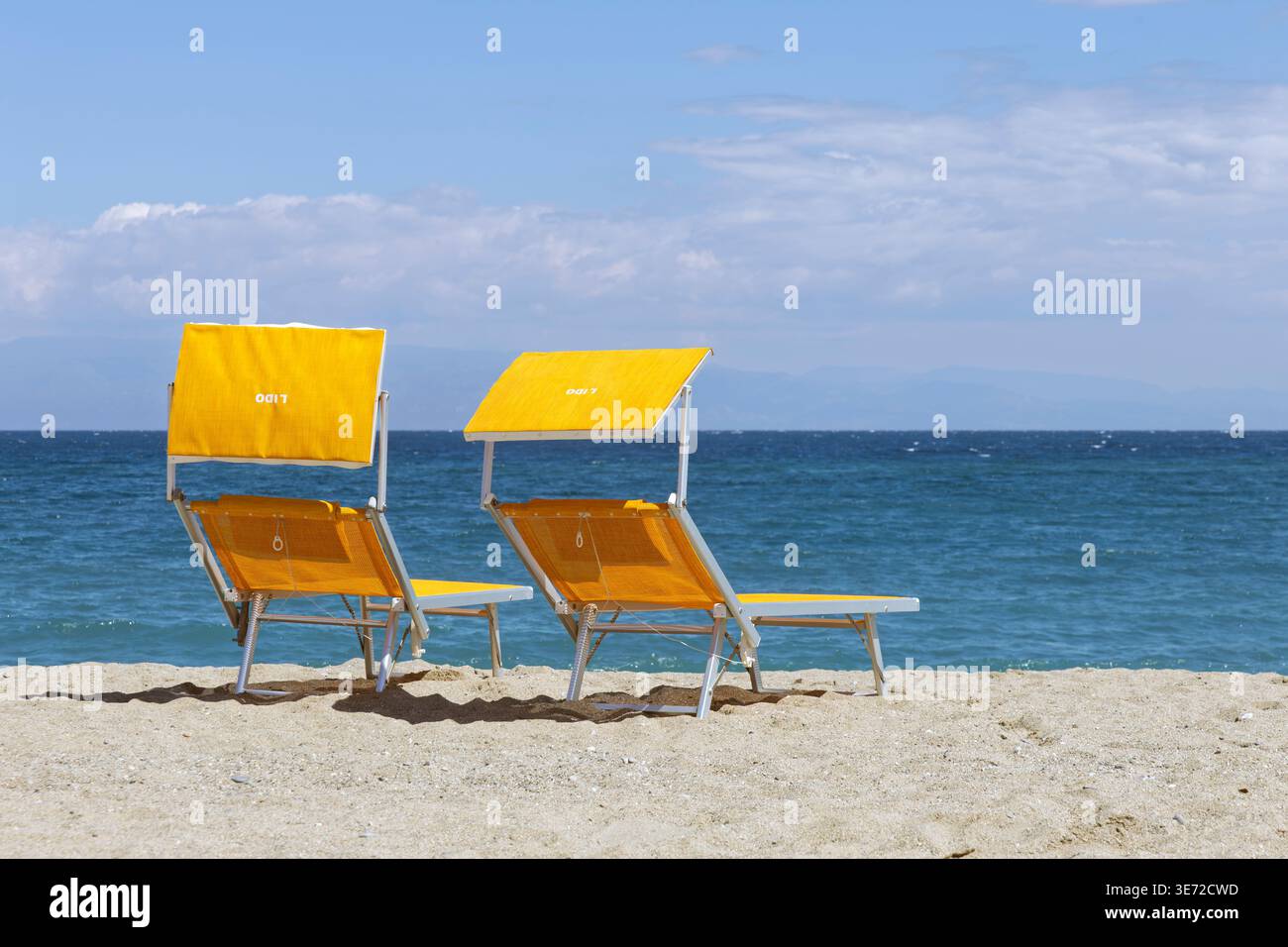 Zwei gelbe Sonnenliegen mit Schatten am Sandstrand von Giardini Naxos, Sizilien, Italien, mit Blick auf das ruhige blaue Wasser des Mittelmeers. Stockfoto