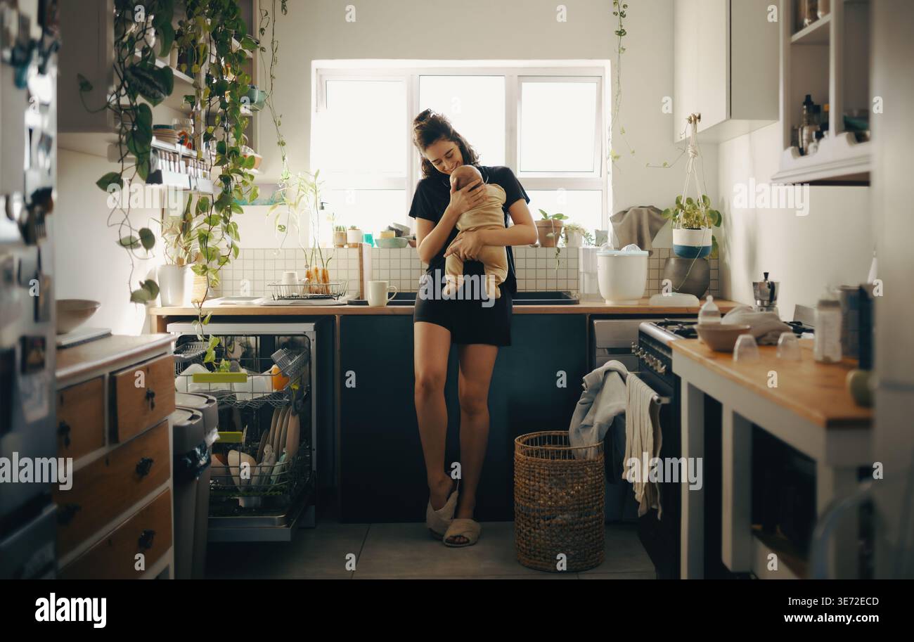 Eine Mutter wiegt ihr Baby in einer sonnendurchfluteten Küche mit Pflanzen und alltäglichen Küchenutensilien. Ein zärtlicher Moment der Pflege und des Heimlebens. Stockfoto