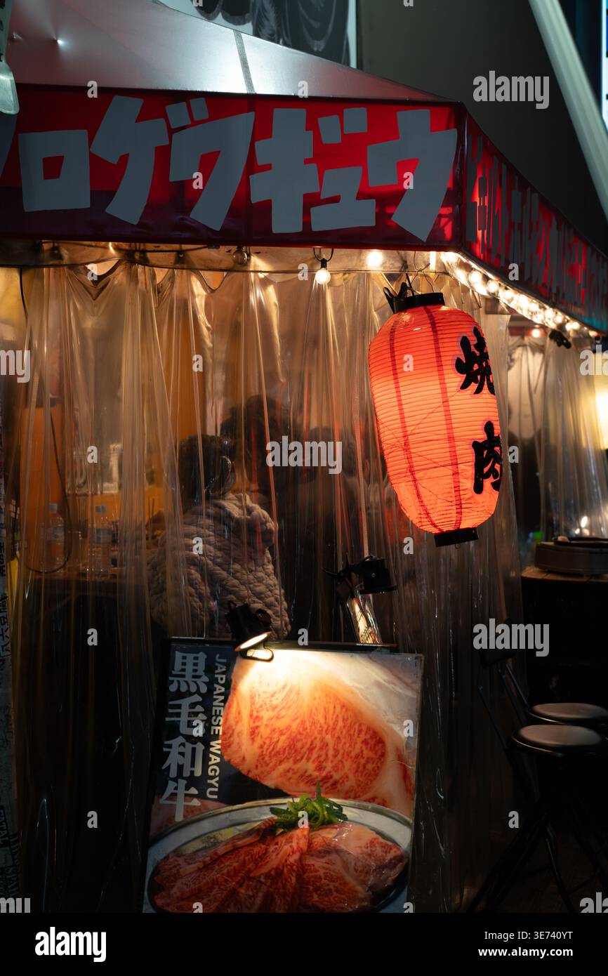 Eine leuchtend rote Laterne hängt vor einem intimen japanischen Yakiniku Wagyu Beef Street Food Stand in Osaka, der von Plastikvorhängen umgeben ist. Stockfoto