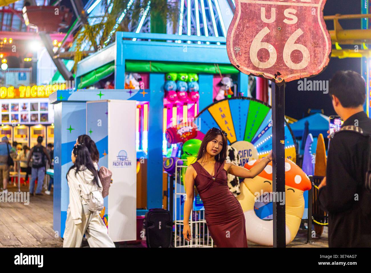 Frau in rotem Kleid posiert mit dem Schild Route 66 am Santa Monica Pier in der Nähe von Los Angeles, Kalifornien, nachts im Pacific Park Vergnügungspark Stockfoto