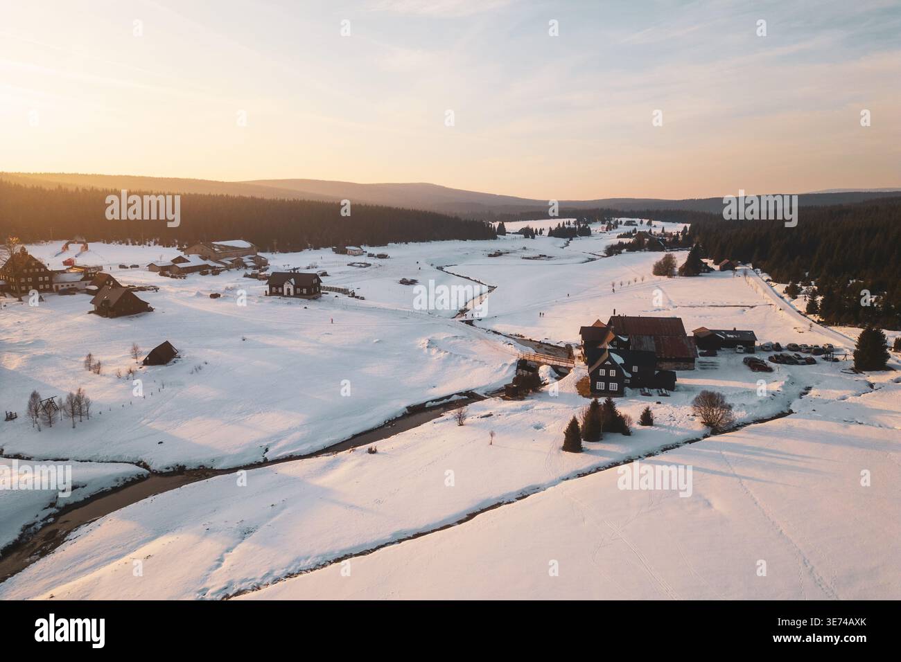 Das Bergdorf Jizerka zeigt im Winter Häuser und schneebedeckte Felder. Die Luftaufnahme fängt die Landschaft mit Bäumen und einem Fluss bei Sonnenuntergang ein. Stockfoto