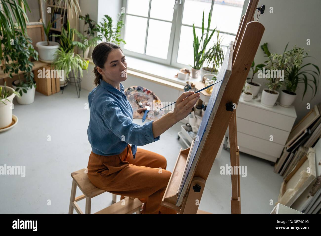 Konzentrierte Malerin zeichnet Bild auf Staffelei mit Palette und Pinsel im modernen Loft. Stockfoto