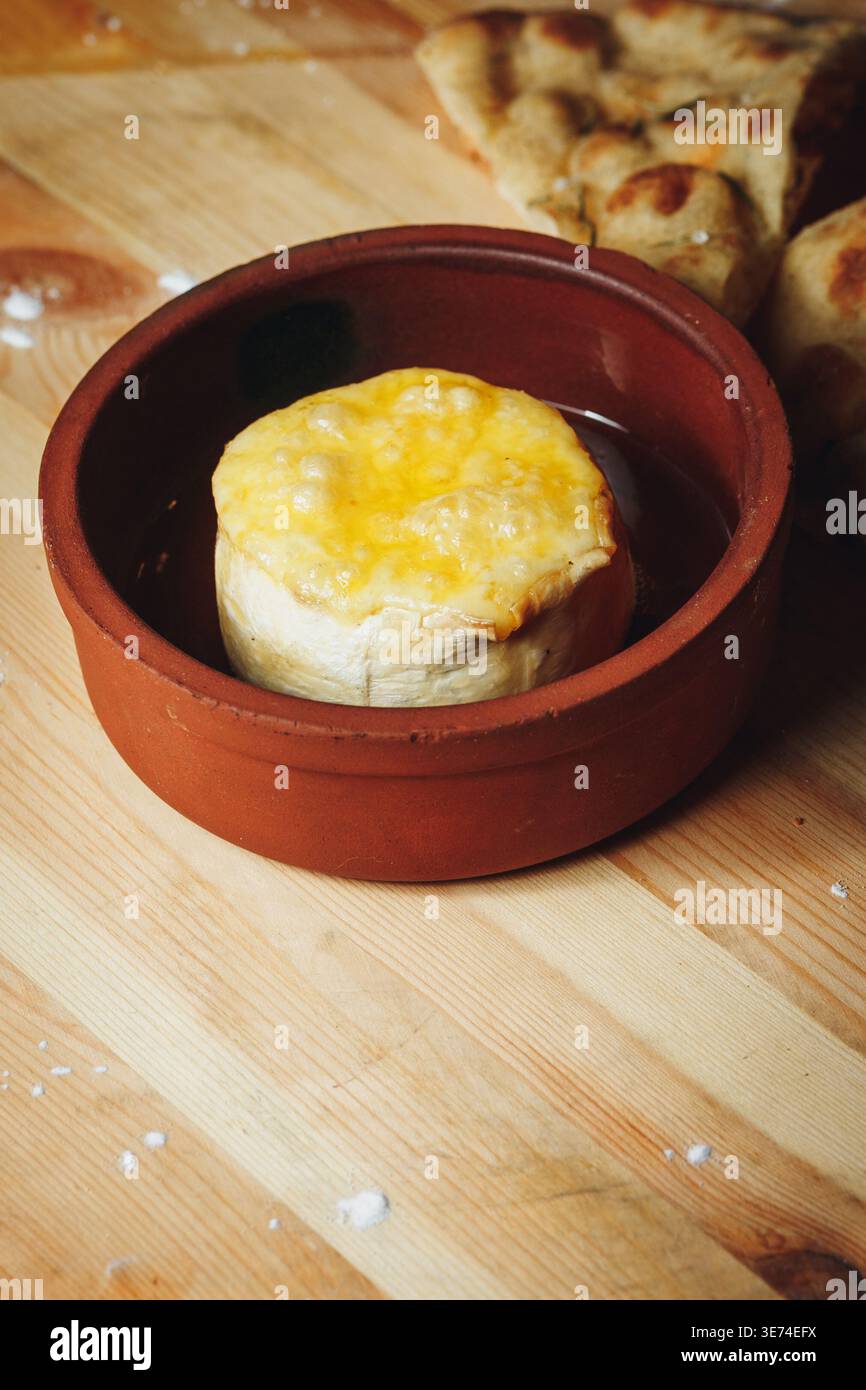 Gebackener Camembert-Käse mit geschmolzener Kruste in einem traditionellen Tontopf, serviert mit Fladenbrot auf einem Holztisch. Stockfoto